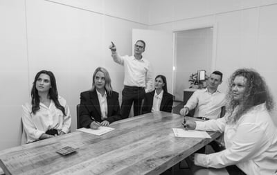 Black & white photo of business meeting: 3 women, 1 man listening, accountant standing and pointing, woman observing, papers & calculator on table.