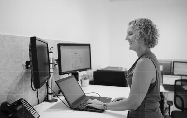 A woman with curly hair is standing at a desk, typing on a laptop. Two monitors are mounted on the wall in front of her, and a phone is on the desk. The setting appears to be a modern office space for a small business focused on cybersecurity, with additional workstations in the background.