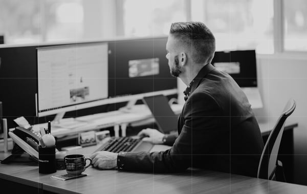 A man in a suit is sitting at a desk, working on his computer with multiple monitors. He has a keyboard and mouse in front of him, a coffee mug on his desk, and appears to be focused on cyber security tasks. The background is blurred, suggesting an office environment in a small business.