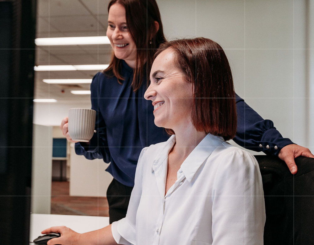 Two business accountants collaborating at a computer, with one holding a coffee mug, discussing financial solutions for clients.