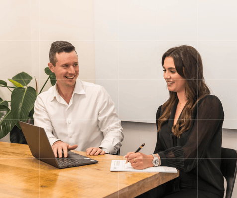 Two people at a table: A smiling male accountant on the left uses a laptop, while a woman on the right writes on a notepad. Background: a plant.