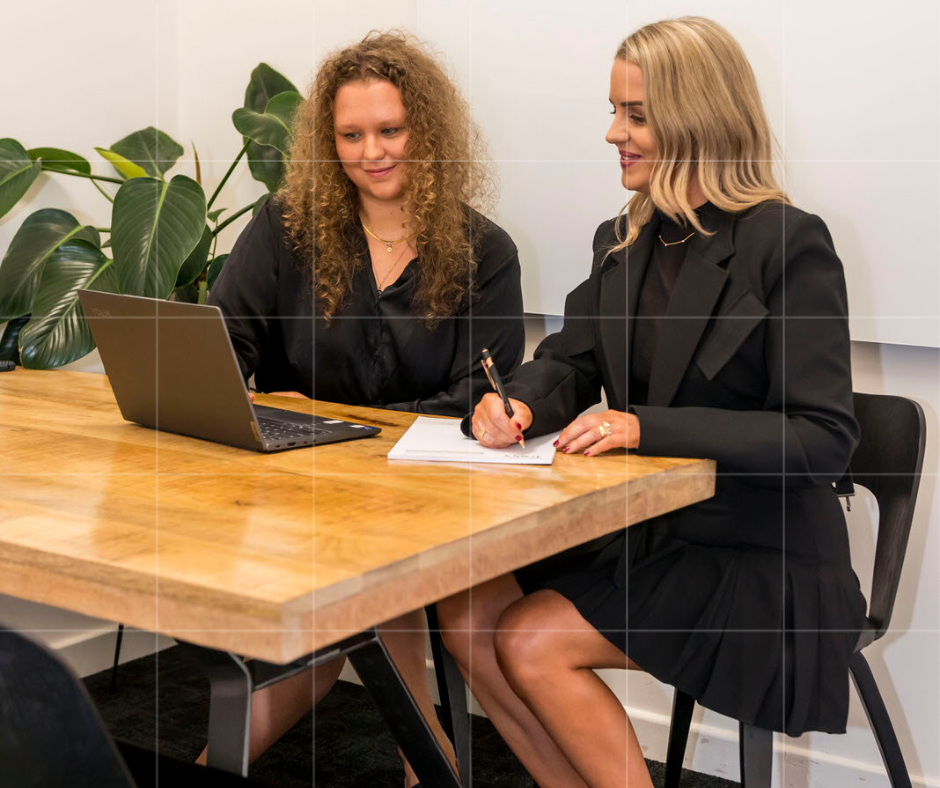 Two women at a wooden table in a modern office. One uses a laptop, the other takes notes on a notepad. A large green plant adds greenery to the space.
