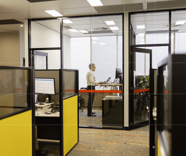 A modern business office space with glass-walled rooms and cubicles, featuring an accountant working at a computer on a standing desk, viewed through the glass. The office has a colorful interior with yellow panels
