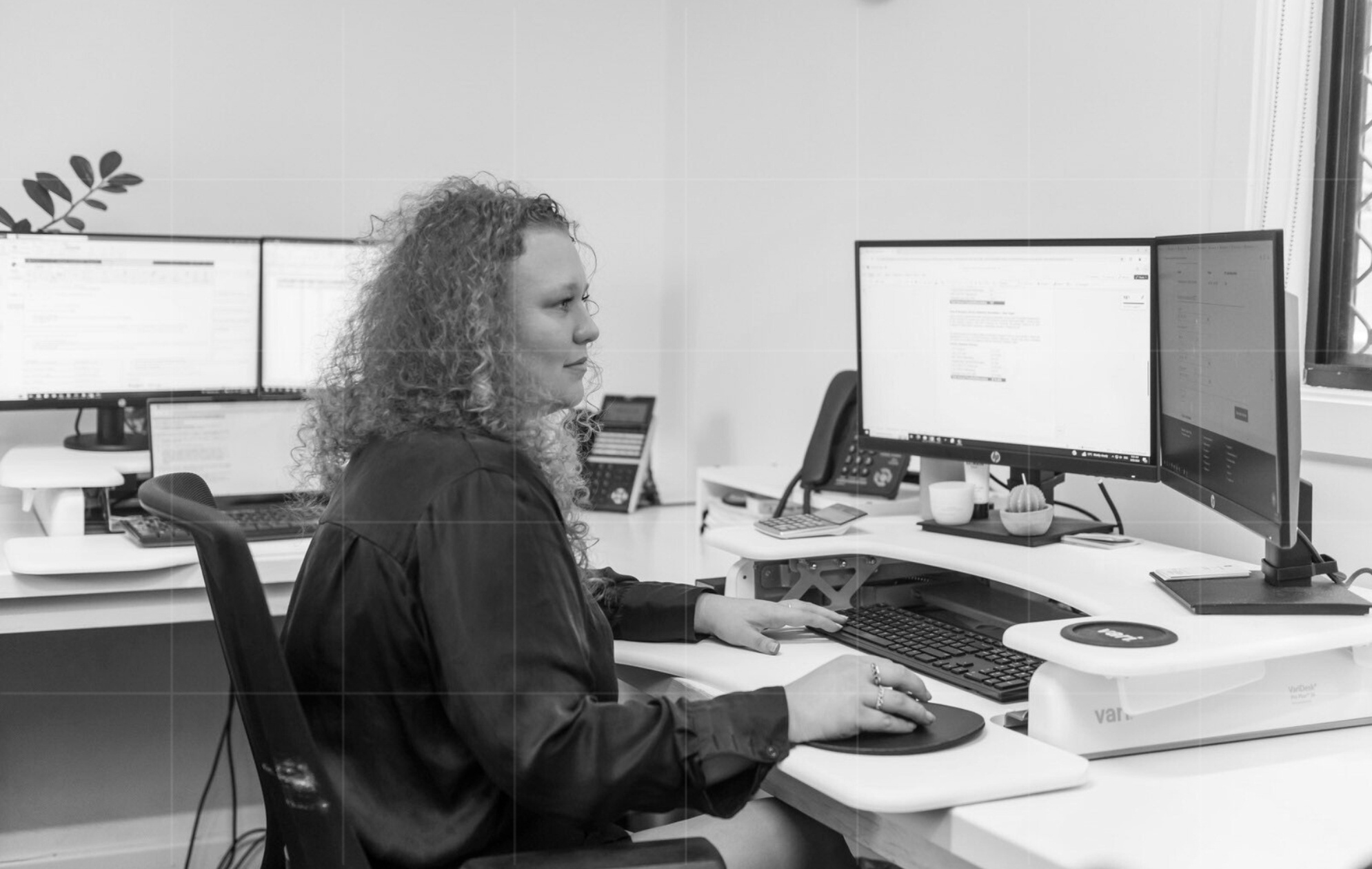 A curly-haired woman in professional attire works at a modern desk with dual monitors in a bright office, surrounded by office supplies and plants.