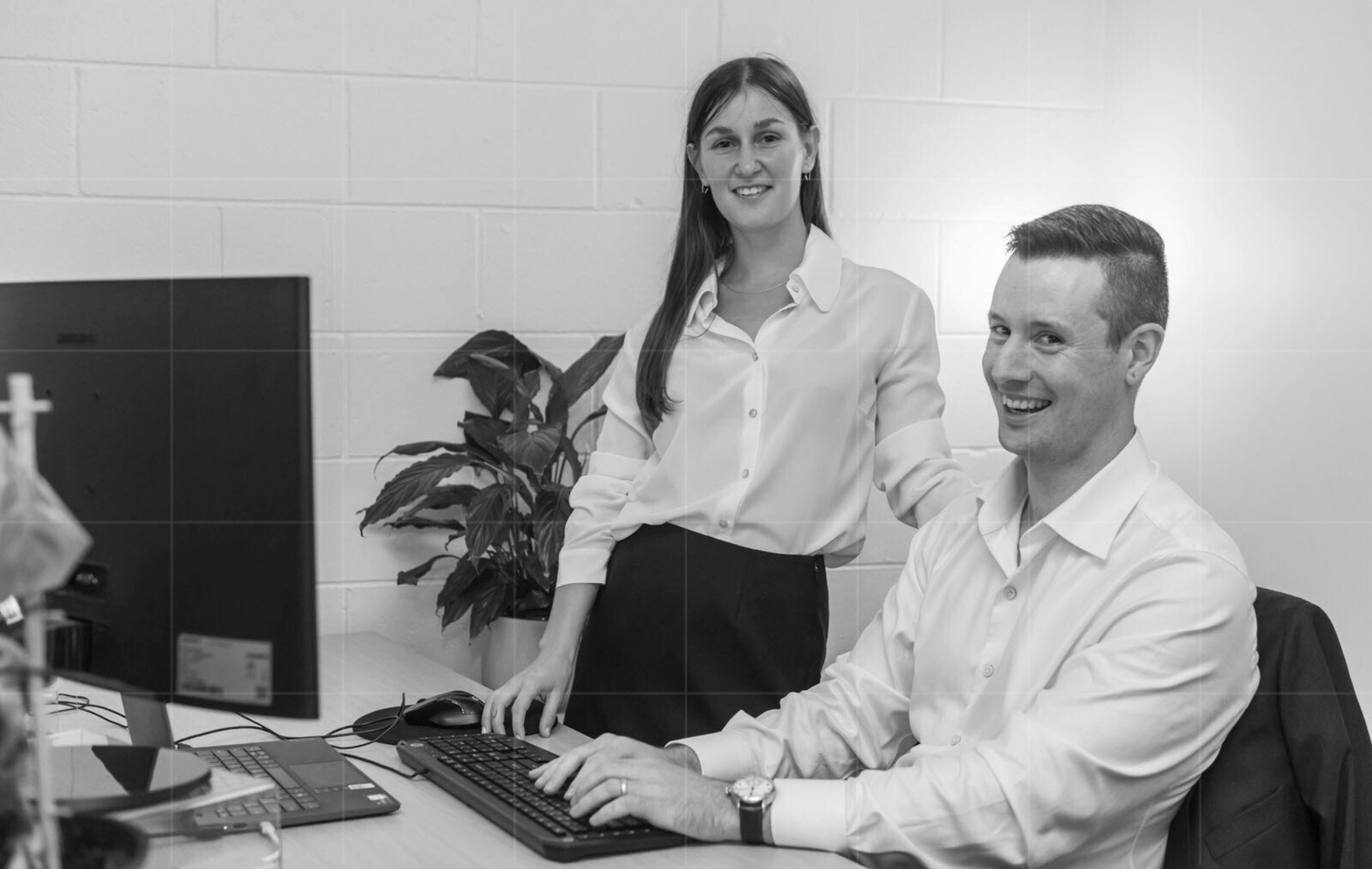 A black-and-white photo of an accountant typing at a desk, smiling at the camera, with a woman beside him, hand on his shoulder, in business attire.