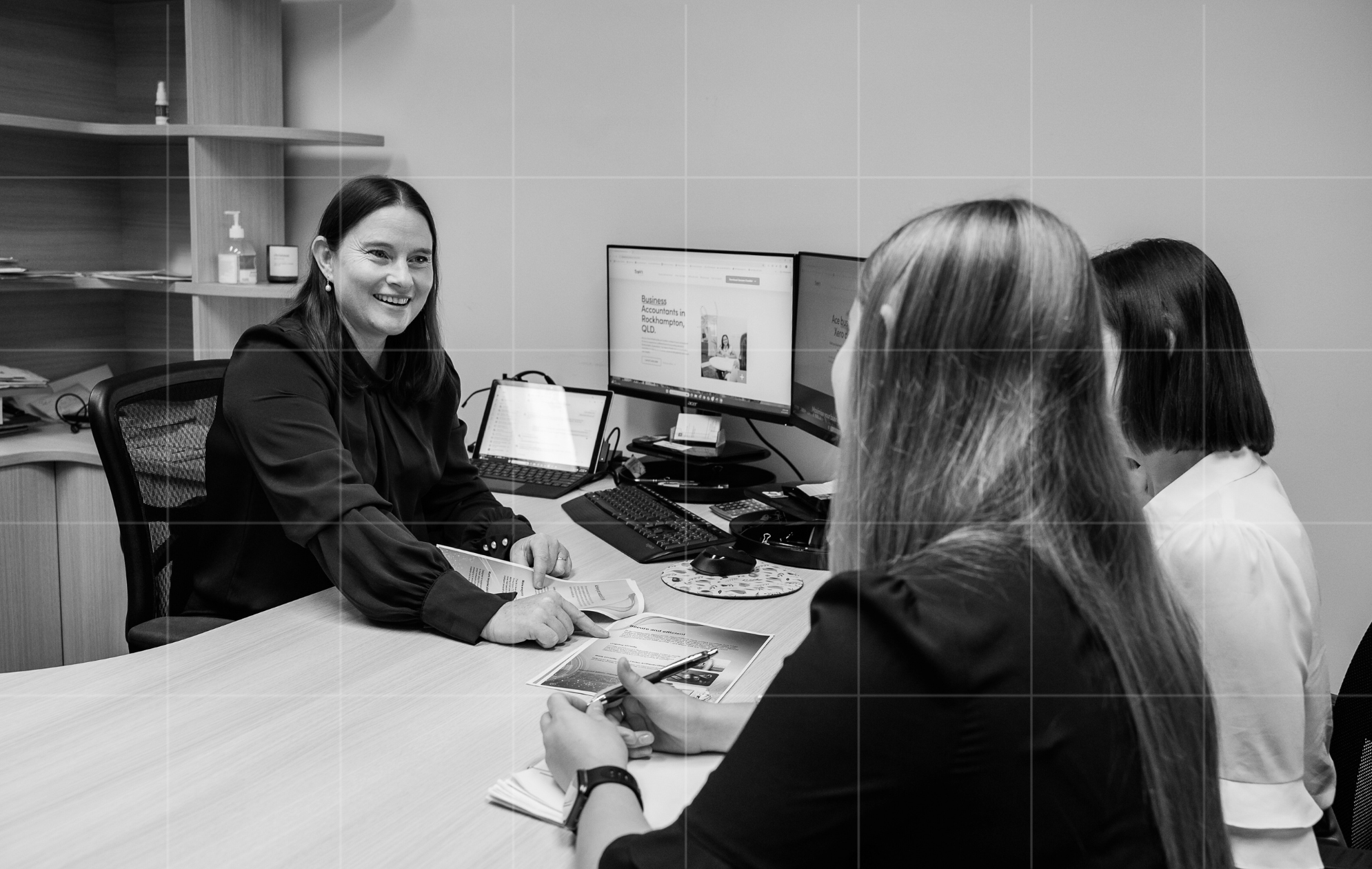 Three businesswomen discussing financial documents in an office, with a website about business accounting displayed on dual monitors - a collaborative session focused on cash flow planning.