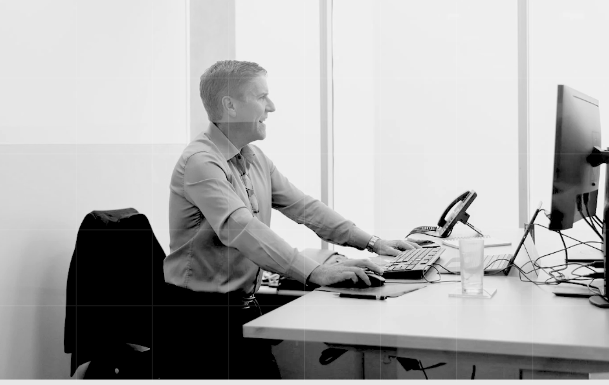 Professional man working at a standing desk in an office environment, smiling and engaged with a desktop computer.