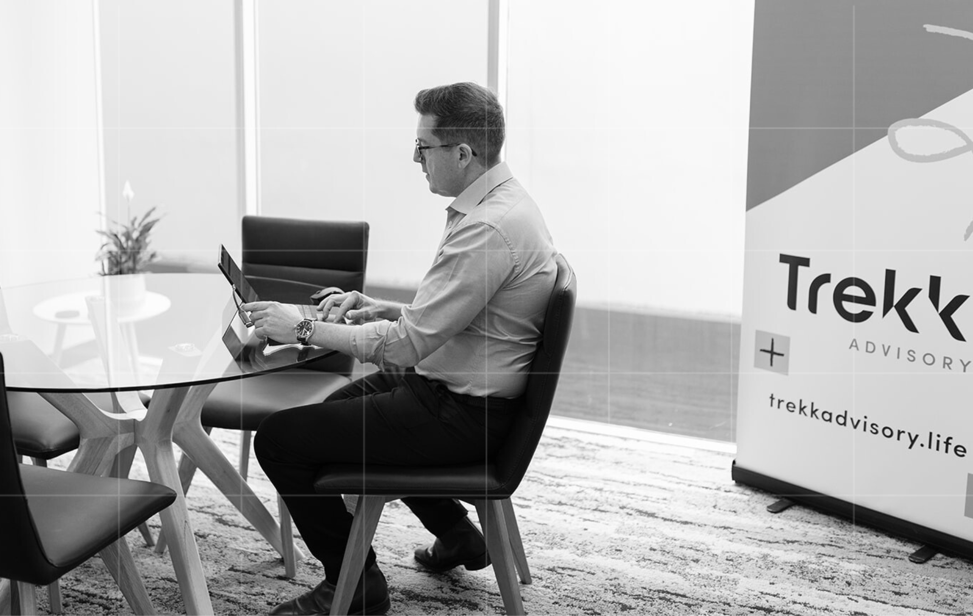  professional man in a business setting, working on his laptop at a glass conference table. A Trekk Advisory banner is visible in the background.