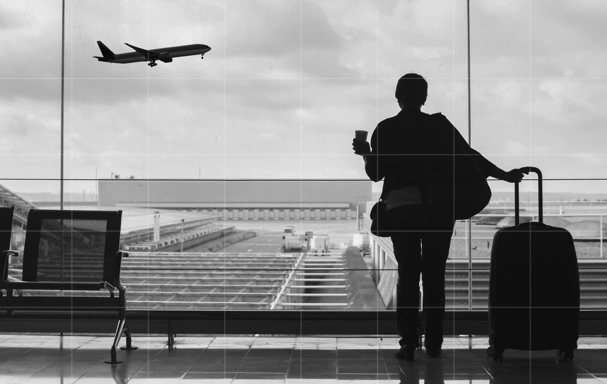 Silhouette of a traveller with a suitcase and coffee cup watching a plane take off from an airport terminal window